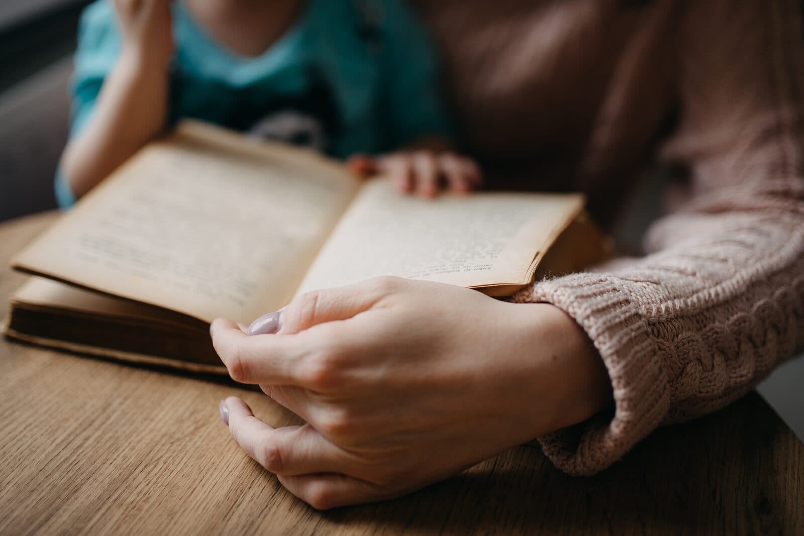 Masada açık duran bir kitap; pembe kazaklı bir yetişkinin eli kitabın önünde, yanında oturan çocuğun eli sayfanın üstünde